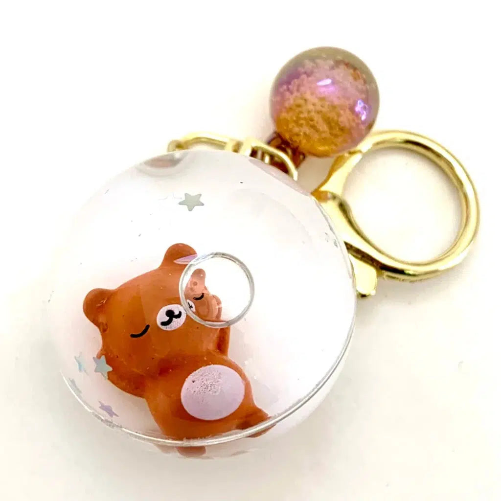 Keychain with a small, smiling brown bear figure inside a clear round globe, attached to a gold keyring and a small, iridescent sphere.