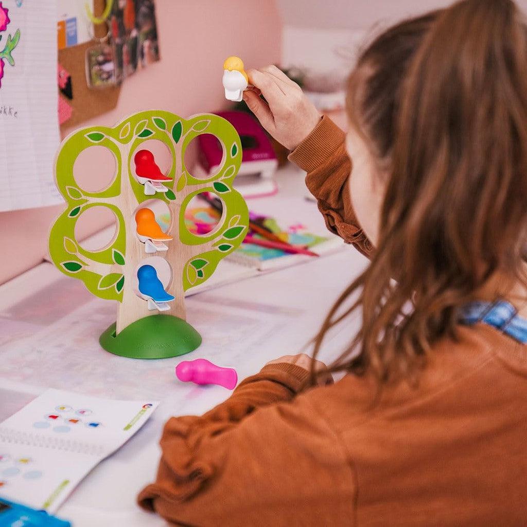 A person plays with a tree-shaped toy featuring colorful birds, reminiscent of stacking games. A book and other items are on the table, inviting thoughtful play like a wooden deduction game from SmartGames.