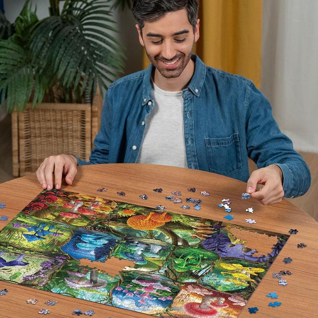 A man assembling a colorful Ravensburger Beautiful Mushrooms jigsaw puzzle at a round table, surrounded by scattered pieces. An inviting scene complemented by a plant and yellow curtain in the background hints at eco-friendly materials used for the puzzle.