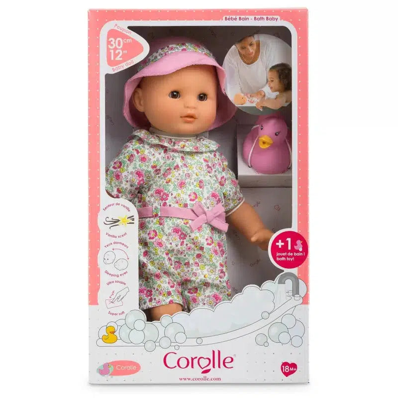 A Corolle bath baby doll in floral outfit and sun hat, packaged in a pink box with illustrations and a toy duck visible.