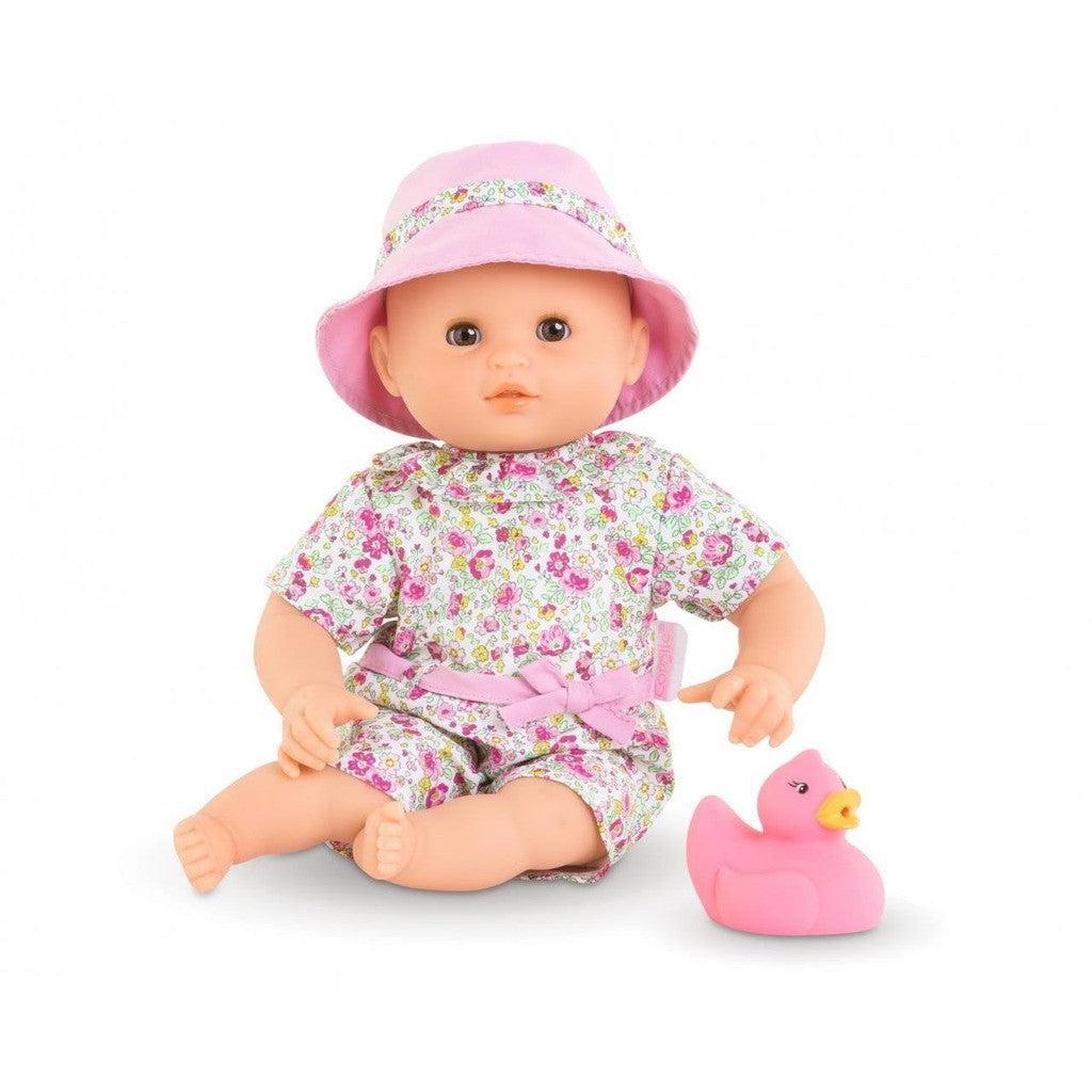 A doll in a floral outfit and pink hat sits beside a pink rubber duck.