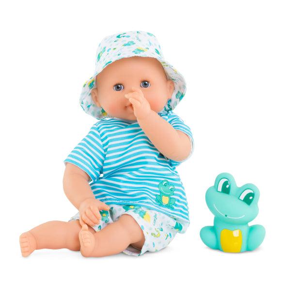 A Corolle baby doll in a striped blue outfit and bucket hat with a frog design sits next to a green plastic frog toy, perfect for water-play fun. The doll curiously places a finger in its mouth, ready for its baby bath adventure.