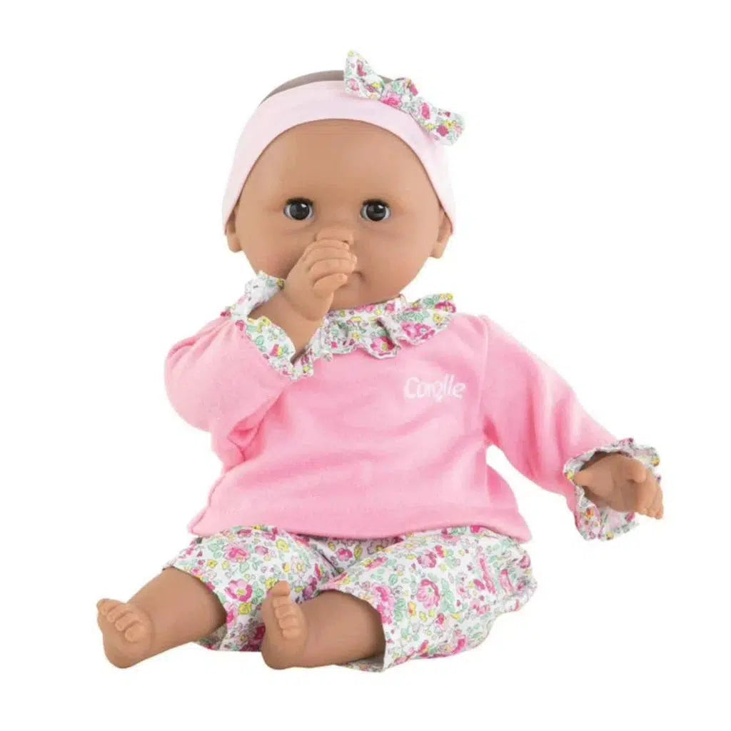 A Corolle doll wearing a pink top with a floral design and matching pants sits with its thumb in its mouth. It has a floral headband adorned with a bow, capturing the charm of Bébé Calin Maria.