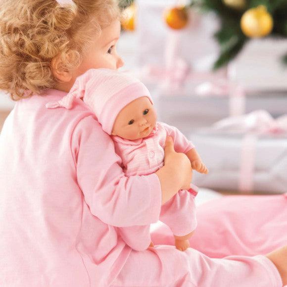 A child with curly hair wearing pink pajamas holds a Calin Charming Pastel baby doll, its soft body dressed in matching attire. They sit in a festive setting, surrounded by the cozy glow of blurred Christmas decorations in the background.