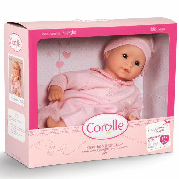 A baby doll in a pink outfit lies in its packaging box, which has a clear front and decorations. The box displays the brand name "Corolle" and the phrase "Création française.