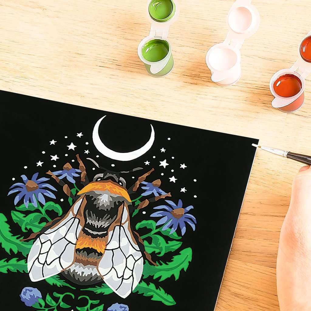 A person is painting a vibrant bee surrounded by flowers and a crescent moon on a black canvas, reminiscent of a Bee Paint by Numbers kit. Open paint pots from Ravensburger CreArt are scattered nearby, adding to the creative ambiance.