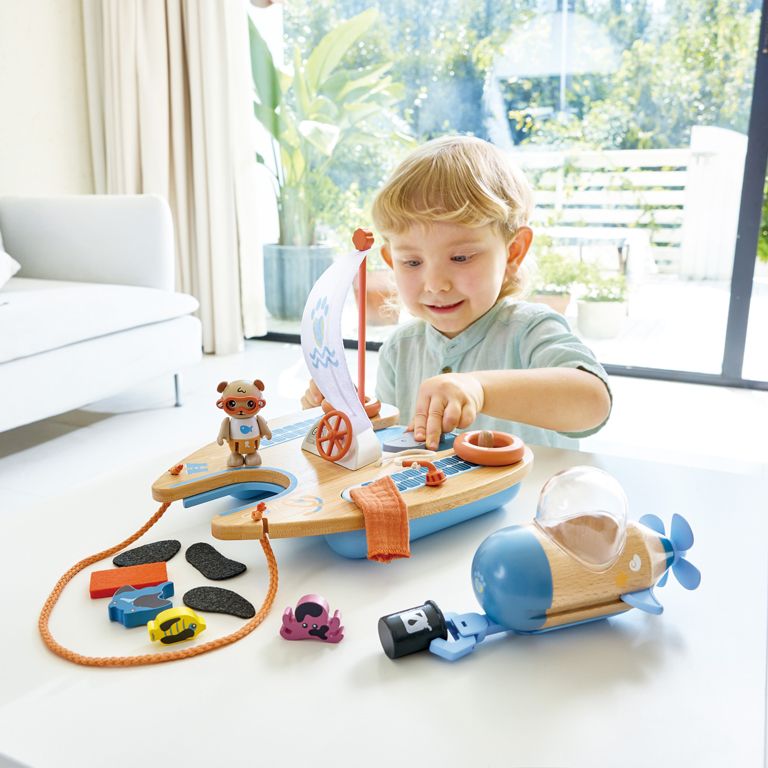 In a bright room with a large window framing the lush greenery outside, a child, dreaming of becoming an eco-conscious sailor, plays with a solar-powered toy boat and airplane set on the table.