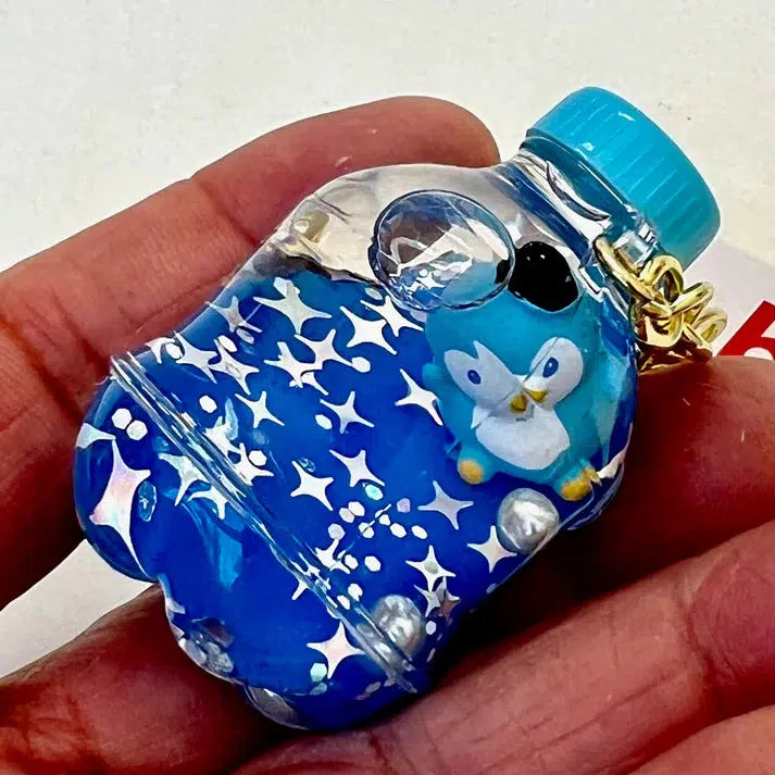 A hand holds a Penguin Floaty Key Charm, showcasing a whimsical clear bottle-shaped keychain adorned with a silly blue penguin design and playful star patterns inside.