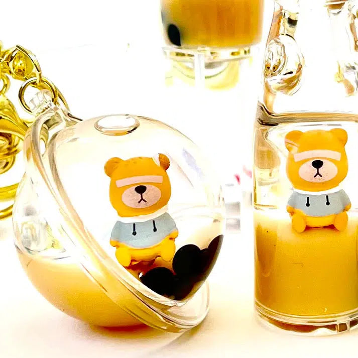 Close-up of two clear containers showcasing small bear figurines, each a delightful Bear Floaty Charm. One bear sports a blue hoodie with a black heart form visible. Background features blurred objects, adding to the charm of these silly animals.