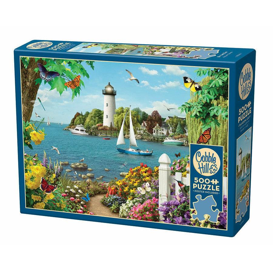 A box of a 500+ piece puzzle showing a vibrant coastal scene with a lighthouse, sailboat, colorful flowers, butterflies, and birds, branded with the Cobble Hill logo.