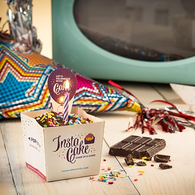 A cake in an InstaCake Celebration Cake Kit box, adorned with sprinkles and a lit candle, surrounded by double chocolate pieces and colorful decorations.