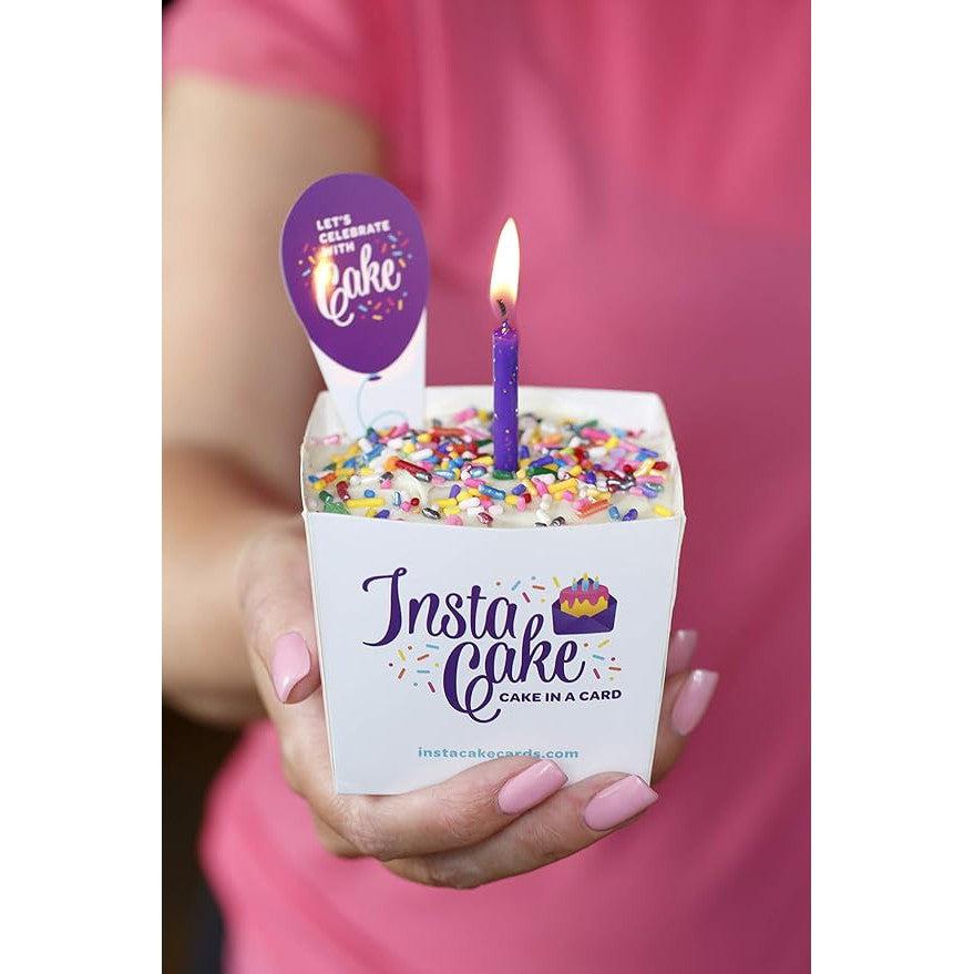 A person holds a small cake in a white box labeled "InstaCake," featuring colorful sprinkles and a lit purple candle, part of the InstaCake Celebration Cake Kit. This delightful treat is perfect for marking special moments, making it an ideal Bakery and Dessert Gift.