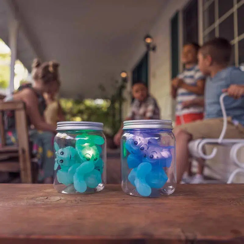 Two jars filled with glowing bear-shaped lights, resembling light-up fireflies, sit on a wooden table, while children enjoy hide-and-seek games on the porch in the background.