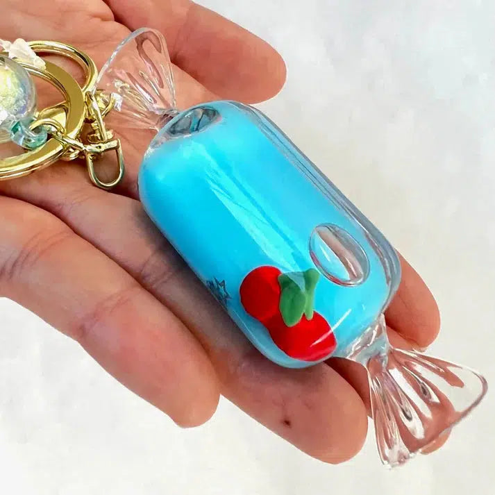 A hand holds a whimsical candy-shaped keychain with a blue filling, reminiscent of cherry candy floaty delights, adorned with a small red and green apple decoration.