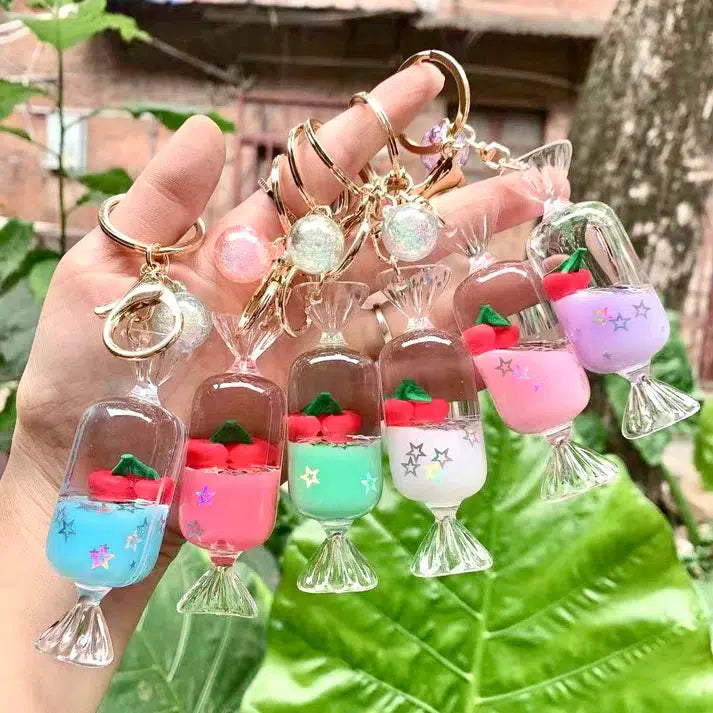 A hand holds colorful cherry candy charm keychains with small charms inside, including fruit and star designs. The playful key ring collection also features silly animals, set against a lush greenery background.