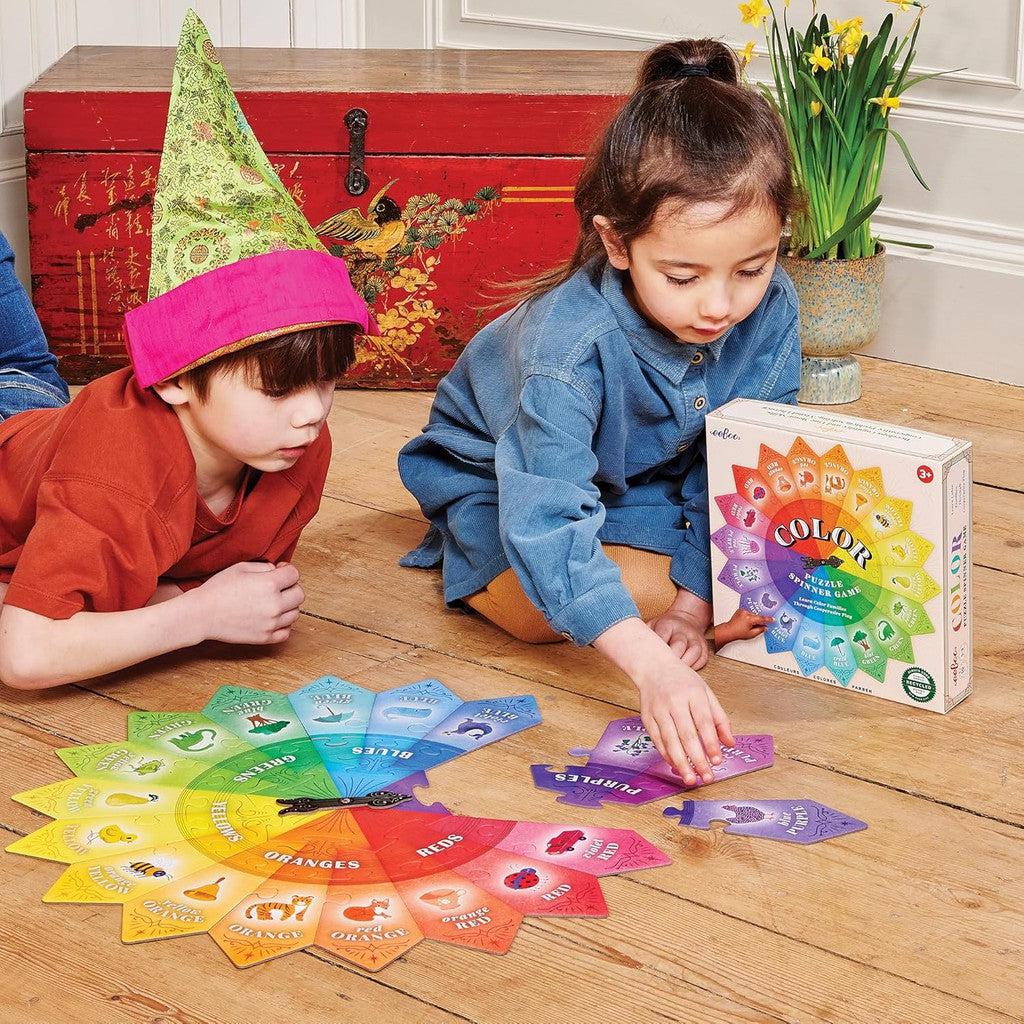 Two children are immersed in a colorful card game on a wooden floor. One, sporting a green and pink pointed hat, eagerly takes their turn with the Color Puzzle Spinner Game. In the background, a red chest and a pot of daffodils add charm to this educational cooperative children's activity for kids ages 3+.