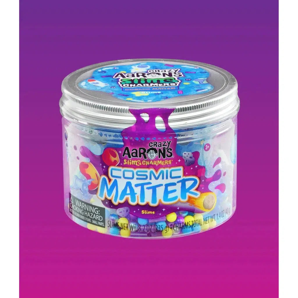 A container of Crazy Aaron's Slime Charmers Cosmic Matter slime, enhanced with colorful beads and tiny space-themed charms, sits against a gradient purple to pink background. The warning label indicates a choking hazard.