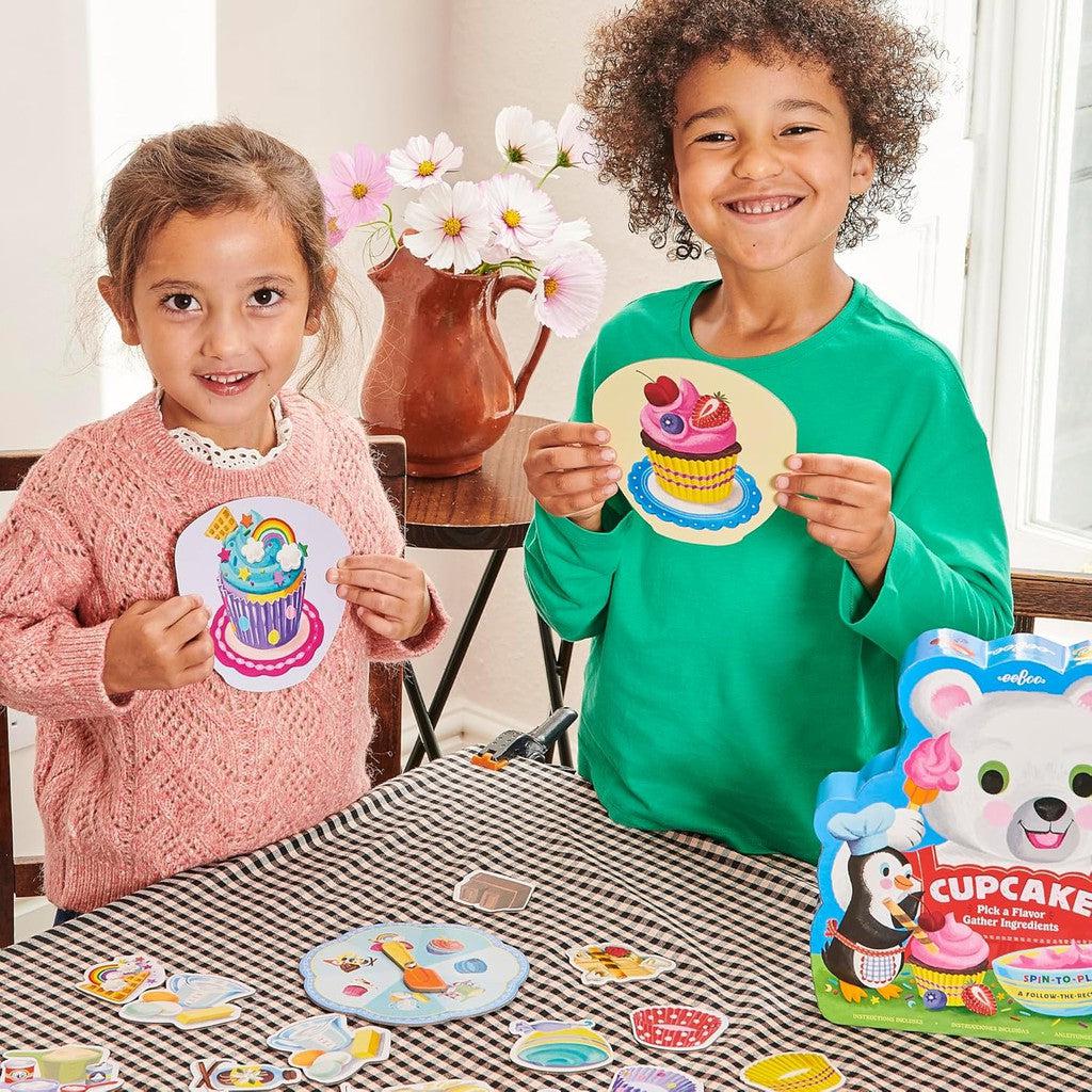 Two preschool children are smiling, holding cupcake-shaped crafts at a table brimming with crafting materials and an eeBoo Cupcake Picnic toy box.