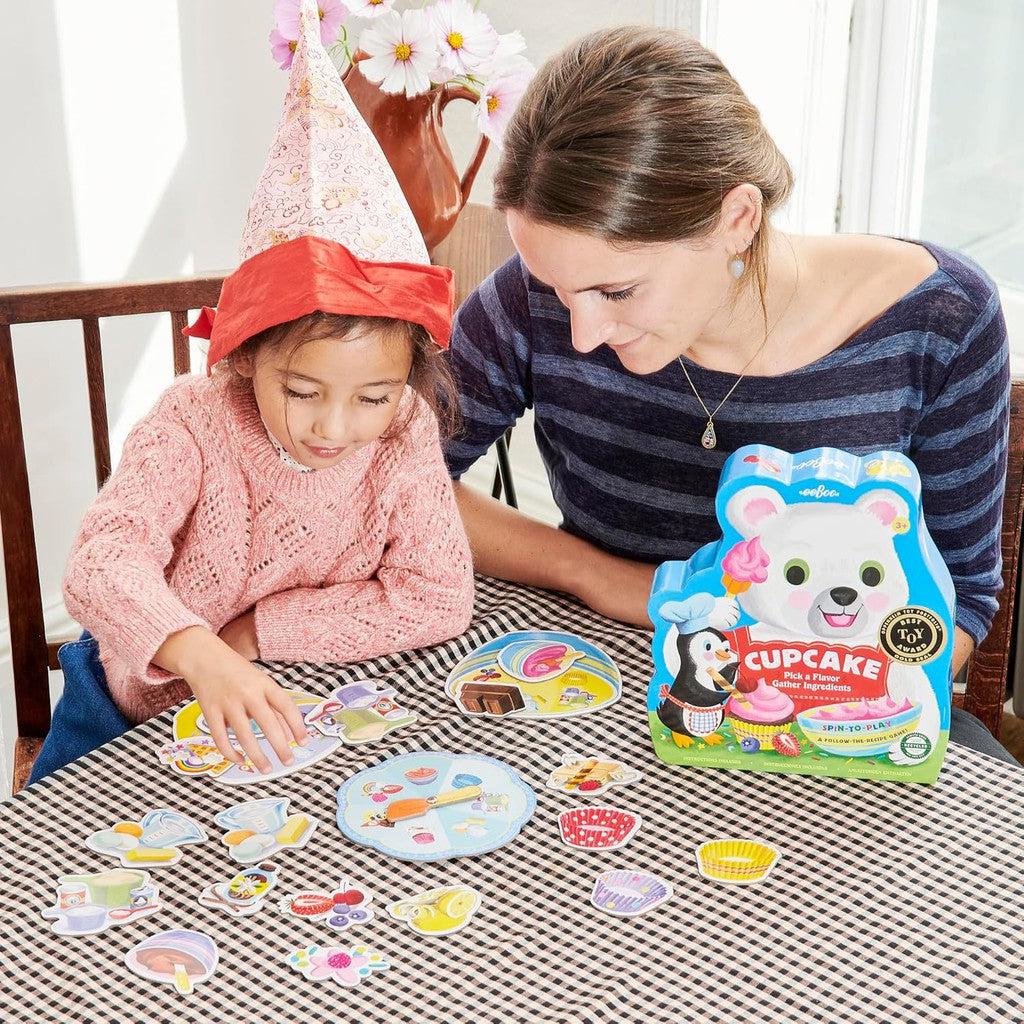 A child in a pink sweater and paper crown joyfully plays a cupcake-shaped spinner game with an adult in a striped shirt. This delightful preschool game creates sweet moments as they match cupcake designs at the table.