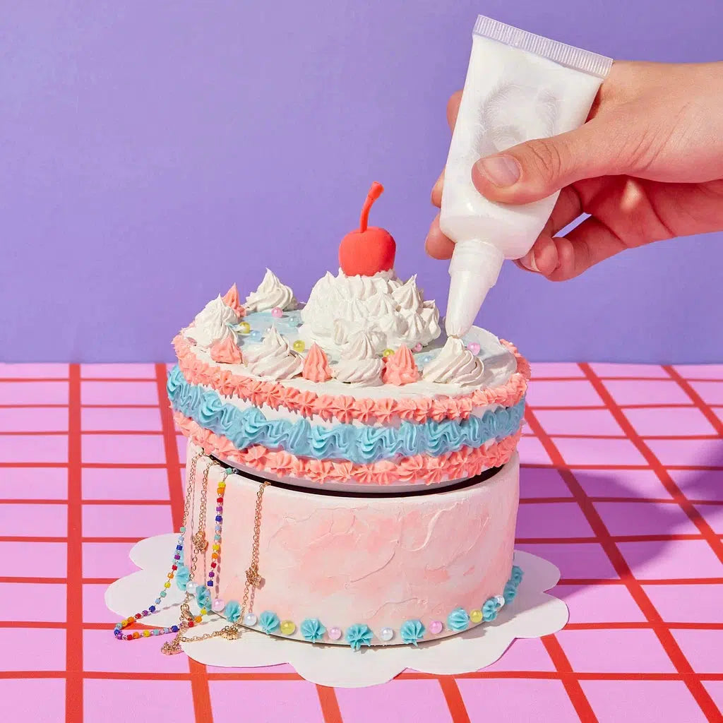 A hand decorates a cake-themed box with white whipped icing clay. The box showcases pink, blue, and white frosting, topped with a red cherry. It's beautifully set against a pink grid-patterned surface with a purple background.