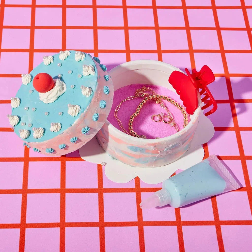 The round blue and white cake-themed box, resembling a ceramic jewelry box, opens to reveal gold chains and rings. Nearby, a pink bow and a blue cream tube rest on the pink grid background, completing the whimsical scene with hints of whipped icing clay detail.