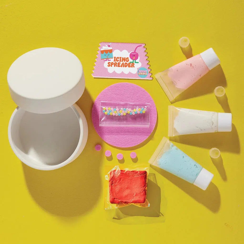 Cake decorating kit with a white container resembling a cake-themed box, an icing spreader card, three icing tubes, red frosting, sprinkles, and small frosting tips on a yellow background.