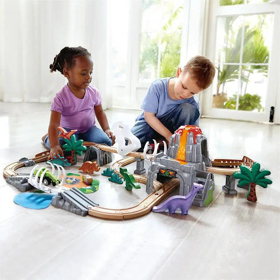 Two children play with a Hape Toys dinosaur-themed train set on a wooden floor. The set from Hape International Inc. includes tracks, a volcano, dinosaur figures, and trees.