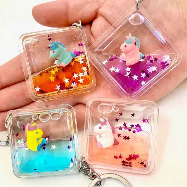 A hand holds four transparent keychains, each with colorful liquid, glitter, and miniature unicorn figures floating inside. These whimsical accessories double as delightful Floating Unicorn Charms, perfect for adding a touch of magic to any key ring.