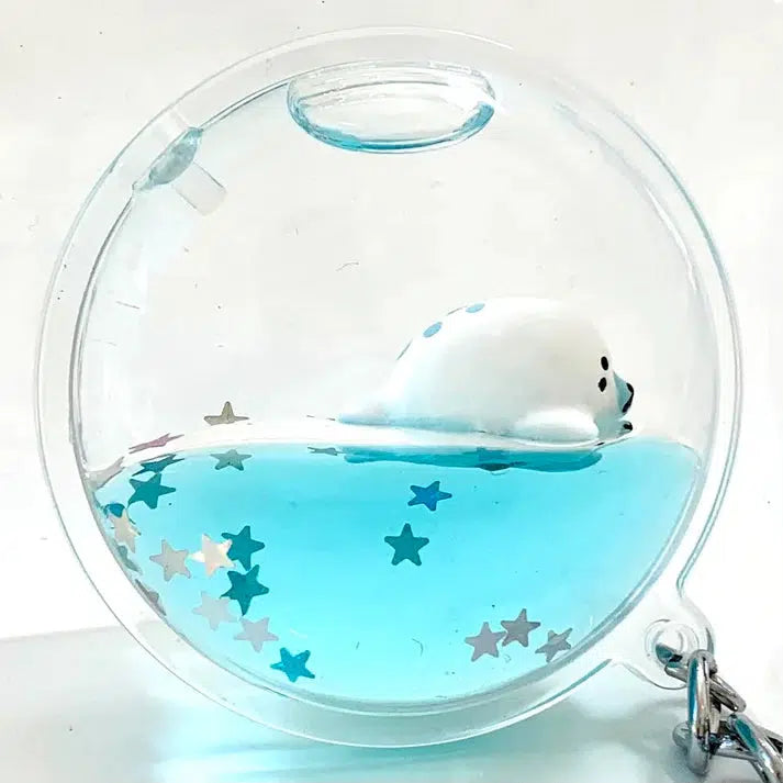 A charming floating sea animal, this small seal toy drifts gracefully in blue liquid inside a clear, round container. Multicolored star-shaped confetti adds a playful touch, transforming it into the perfect floating sea charm for your key ring.