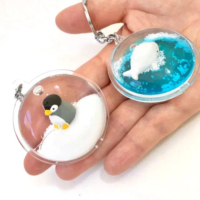 Two small, round keychains held in a hand, featuring silly animals—a penguin in snow and a white seal in blue water. These charming key rings capture whimsy perfectly.