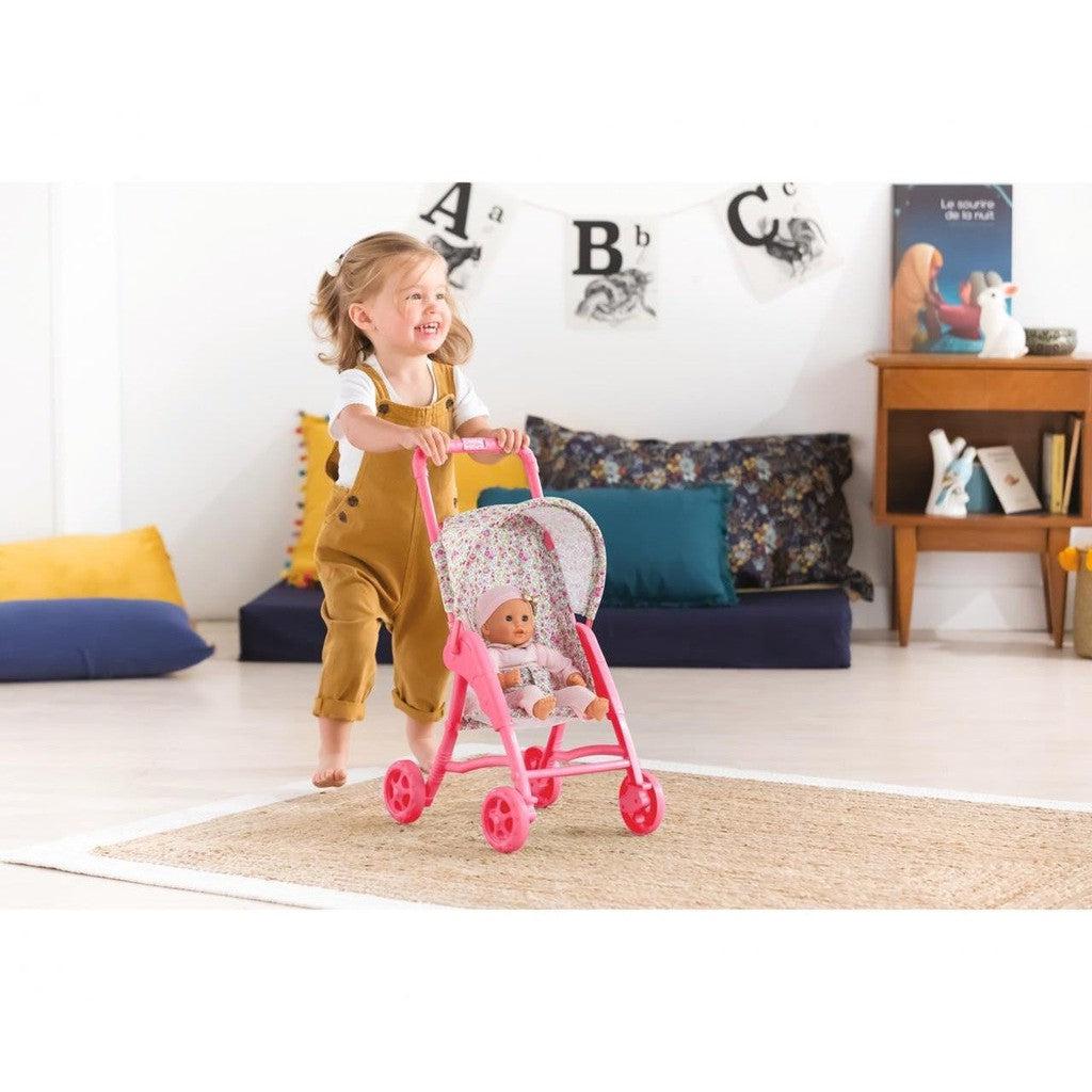 A young child pushes a doll in a pink stroller across a rug in a living room.