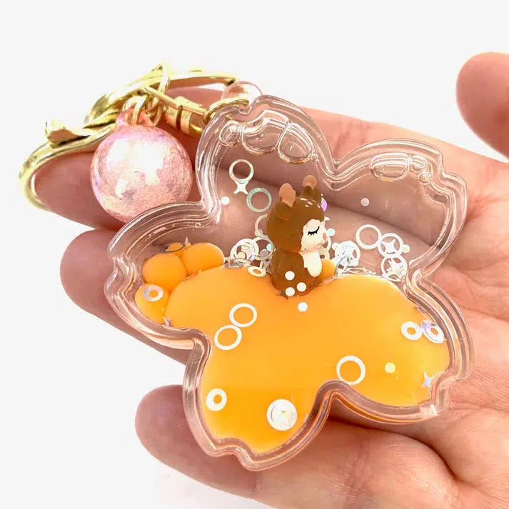 A clear star-shaped keychain with an orange base holds a small brown toy figurine and decorative sequins, shown resting on a person's palm. A shiny pink bead is attached to this whimsical key charm.