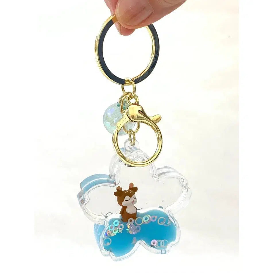 A hand holds a key charm shaped like a transparent whale from the Silly Animals collection, featuring a small toy figurine inside. The keychain contains blue liquid and floating glitter.