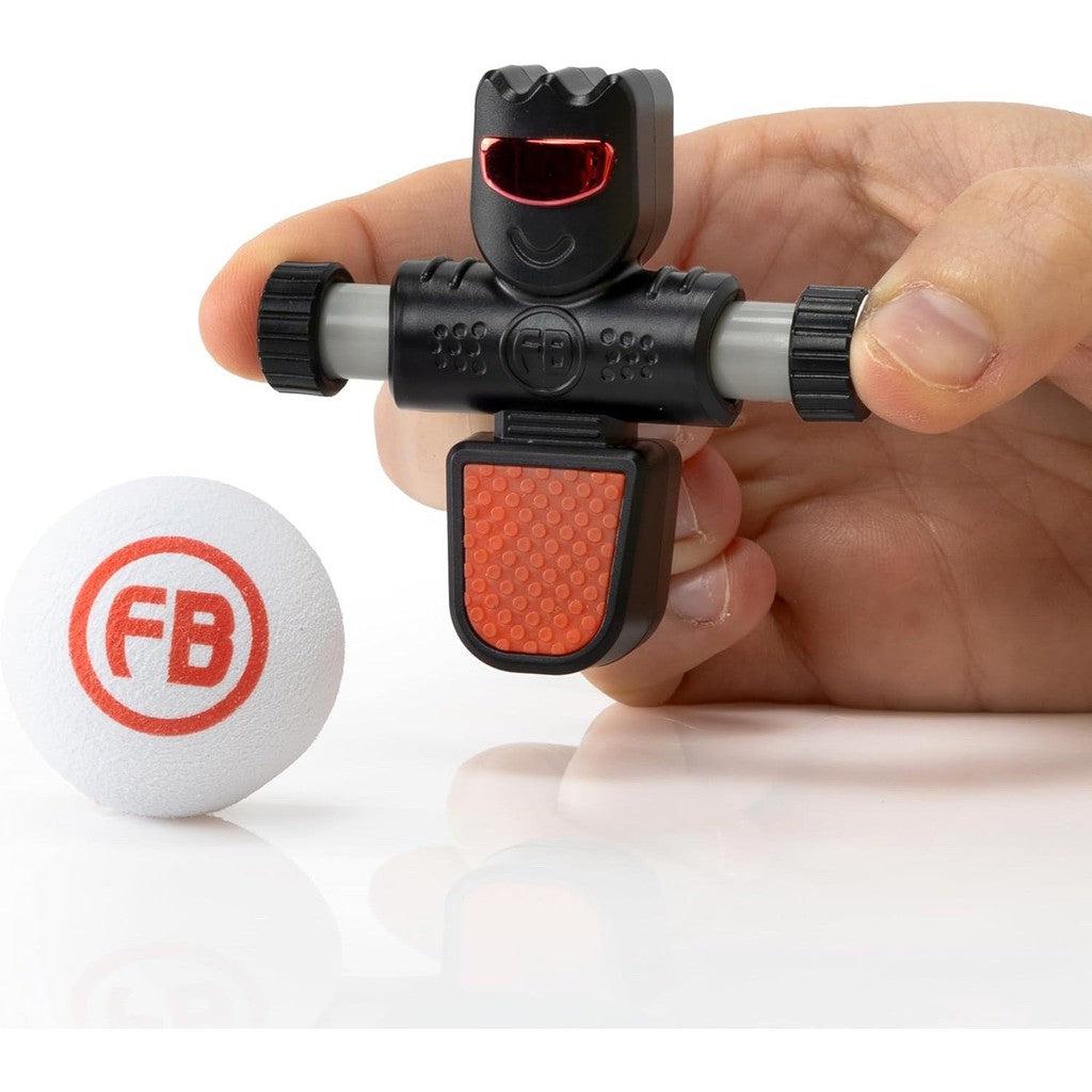 A hand holds a small black toy with a red mouth and chest, part of the Foosbots Series 3 collection. It's next to a white ball labeled "FB" on a white surface, perfect for those who enjoy Toys &amp; Games during Active Play sessions.