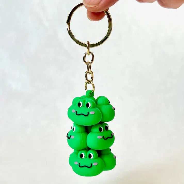 A hand holds a Frog Family Key Charm featuring stacked green frog faces, each with cartoonish eyes and a smiling expression. This adorable toy figurine captures the essence of silly animals in the cutest way possible.