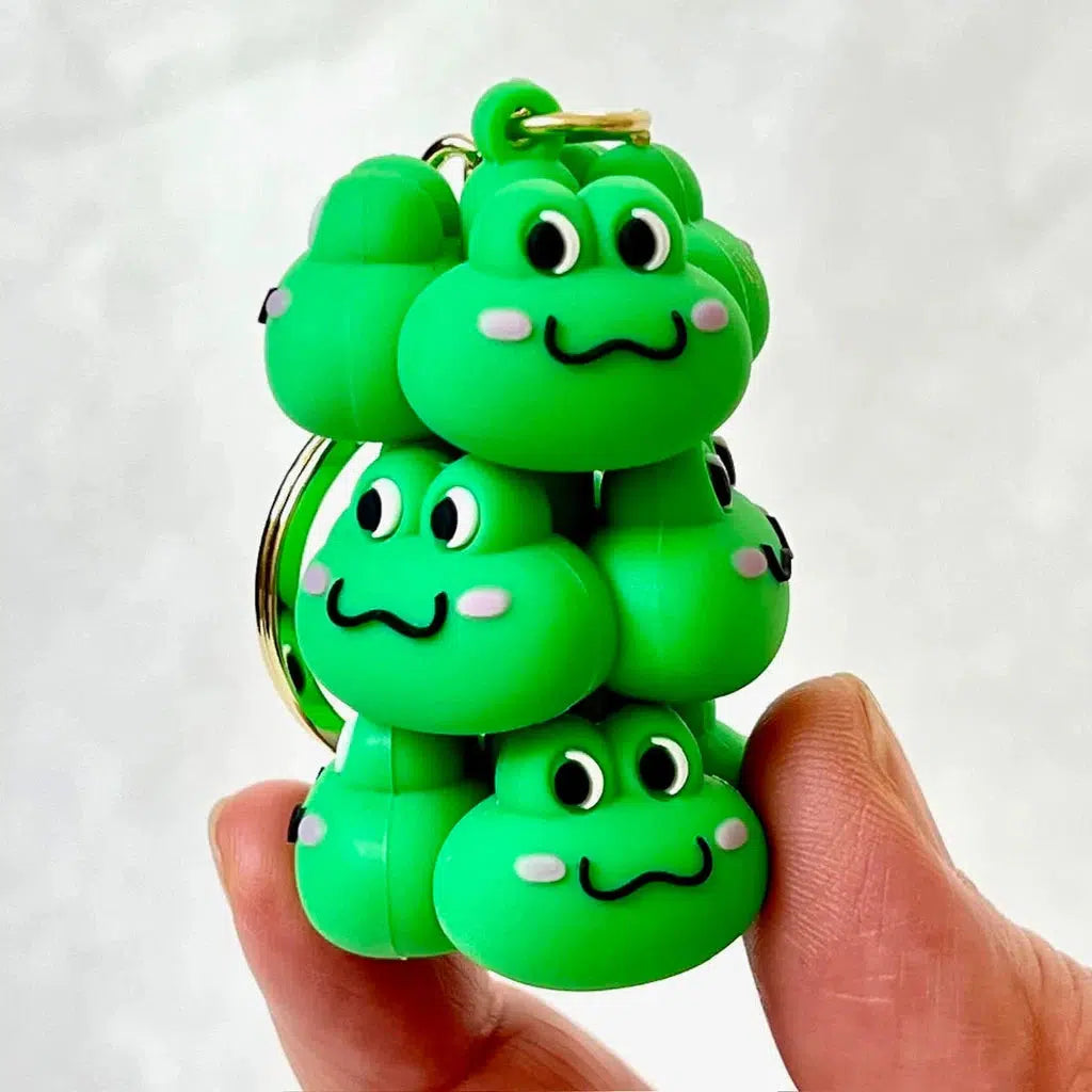 A person holds a Frog Family Key Charm with multiple stacked green frog faces, each grinning wide. This playful charm makes the perfect Backpack Accessory for those who adore Silly Animals Charms.