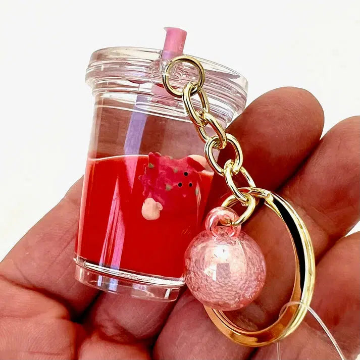 A hand holds a Fruit Boba Key Charm, featuring a cup, straw, and pink decorative element on a gold chain.