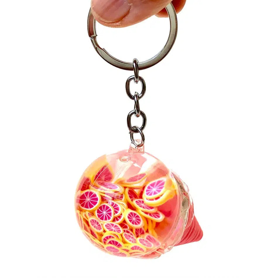 A hand holds a key charm featuring a transparent sphere with vibrant pink and orange citrus slice designs, playfully reminiscent of fruit ice cream on a sunny day.