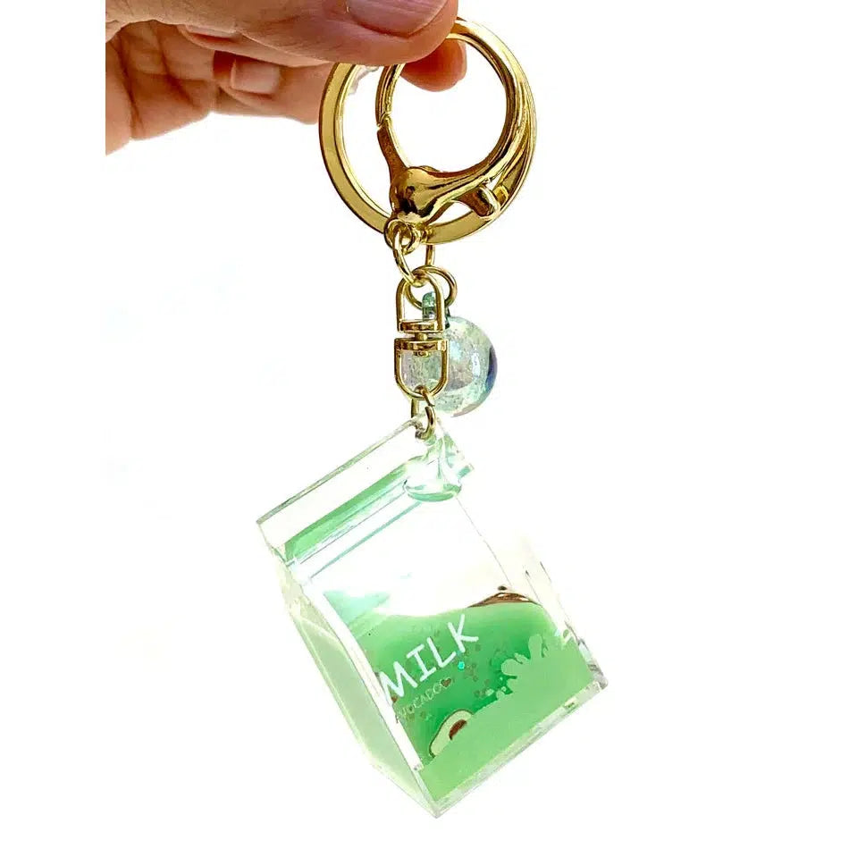 A hand holds a charming keychain with a small, clear milk carton design, now featuring green liquid and a floating sphere, attached to a gold ring and clip. This delightful key charm adds a quirky touch to any accessory collection.