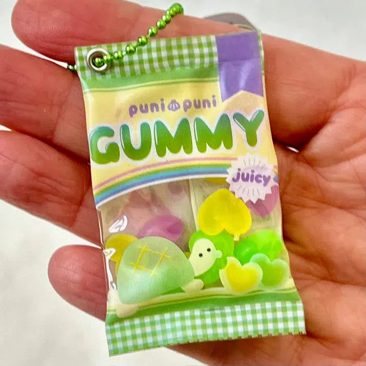 A hand holds a small Gummy Key Charm designed like a packet of gummy candy, featuring colorful fruit shapes and the playful "Puni Puni Gummy" text, offering assorted designs including silly animals.