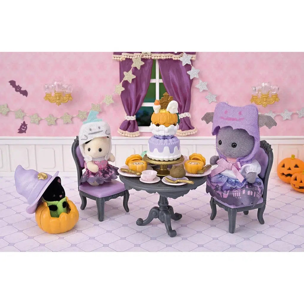 Two calico critter cats in Halloween costumes sit at a table with a decorated cake. A black toy cat in a pumpkin costume is nearby. The room is decorated with stars and bats.