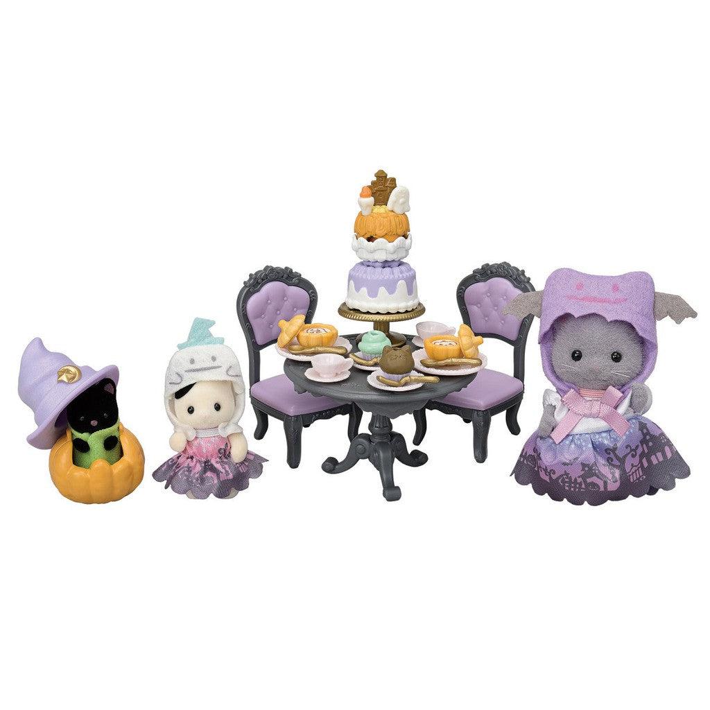 Three Calico Critters dressed in Halloween-themed clothes are gathered around a table set with a cake and tea. The table is surrounded by chairs, and one toy is inside a pumpkin.