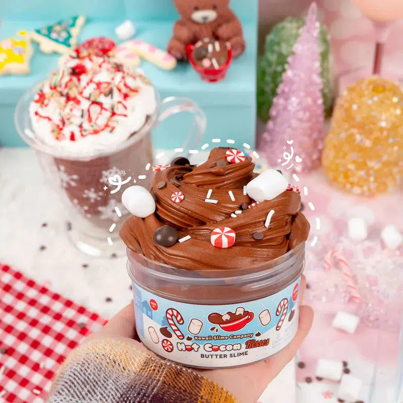 A jar of chocolate-themed butter slime adorned with marshmallows and candy pieces is held in front of festive holiday decor, including a mug of hot chocolate and colorful Christmas trees.