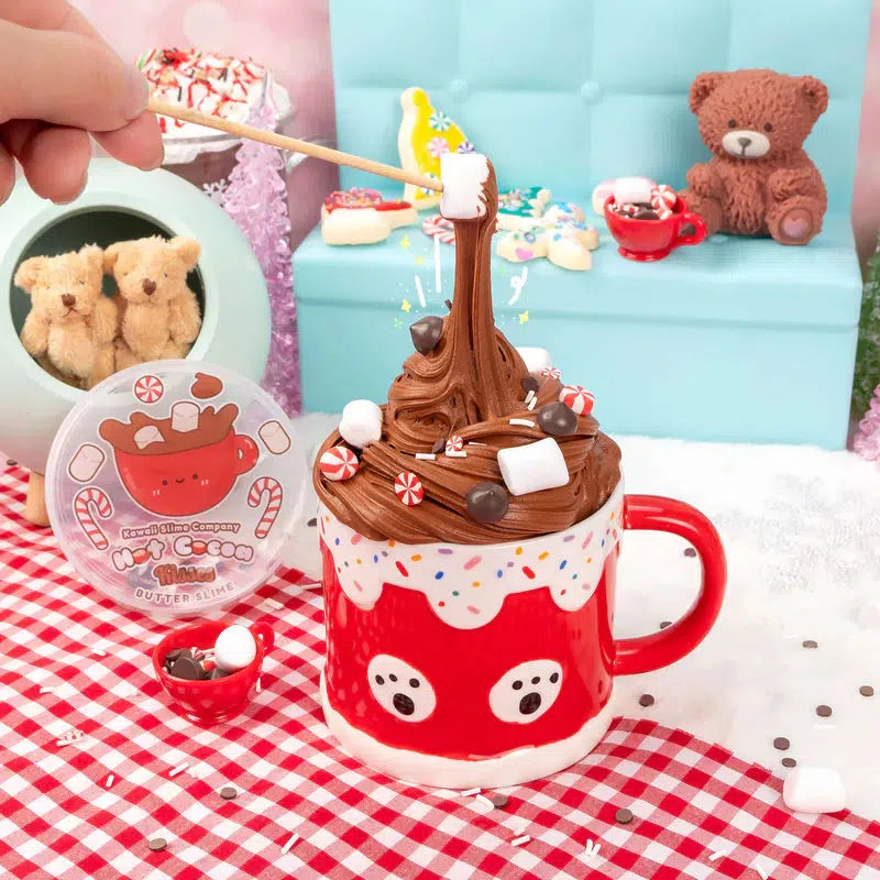 A hand adds small marshmallows to a cup topped with chocolate frosting. The scene includes a red cup with a festive design, and plush bears in the background on a blue sofa.