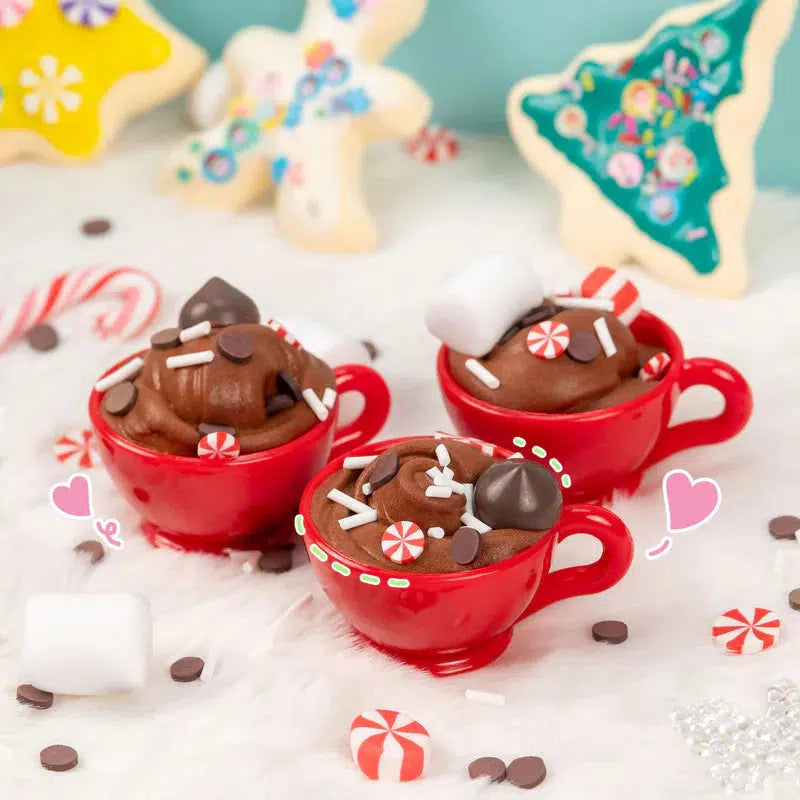 Three red mugs filled with chocolate dessert-looking slime, topped with marshmallows and candy, surrounded by decorated cookies and peppermint candies.