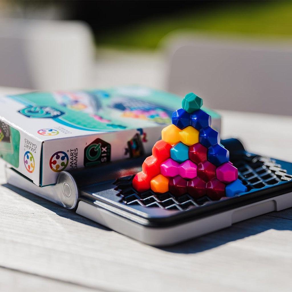 A brain game featuring colorful hexagonal pieces stacked in a pyramid shape sits on a wooden table, its box labeled SmartGames IQ Six Pro nearby. Perfect for travel games enthusiasts, this engaging puzzle promises hours of fun and mental stimulation.