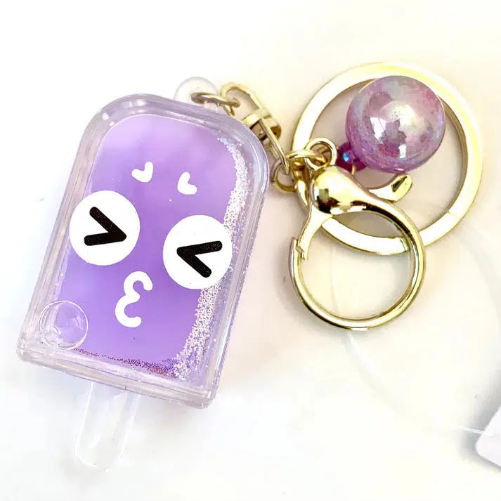 A key ring with a delightful purple popsicle face charm sporting heart eyes, attached to a gold clasp, and accompanied by a pearl-like bead.