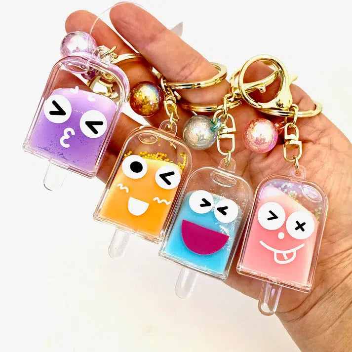 A hand holds four keychains shaped like popsicles with silly cartoon faces in purple, orange, blue, and pink hues, adorned with gold key rings and decorative beads. These delightful toy figurines add a whimsical touch to any collection.