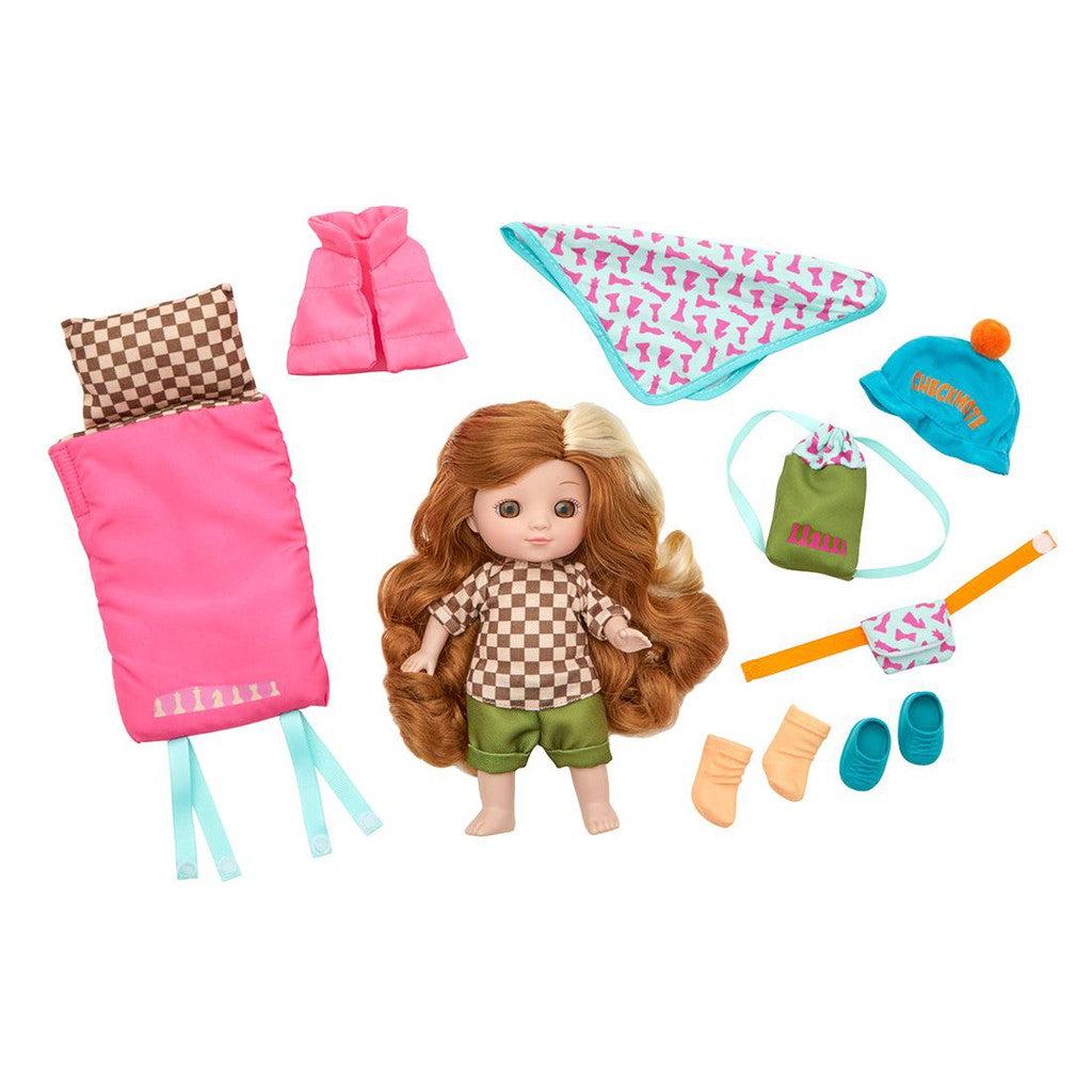 This charming doll, perfect for camping adventures, boasts long brown hair and wears a checkered top with green shorts. She's surrounded by outfits and accessories like a pink sleeping bag, various outfits, a hat, shoes, socks, and a bandana.