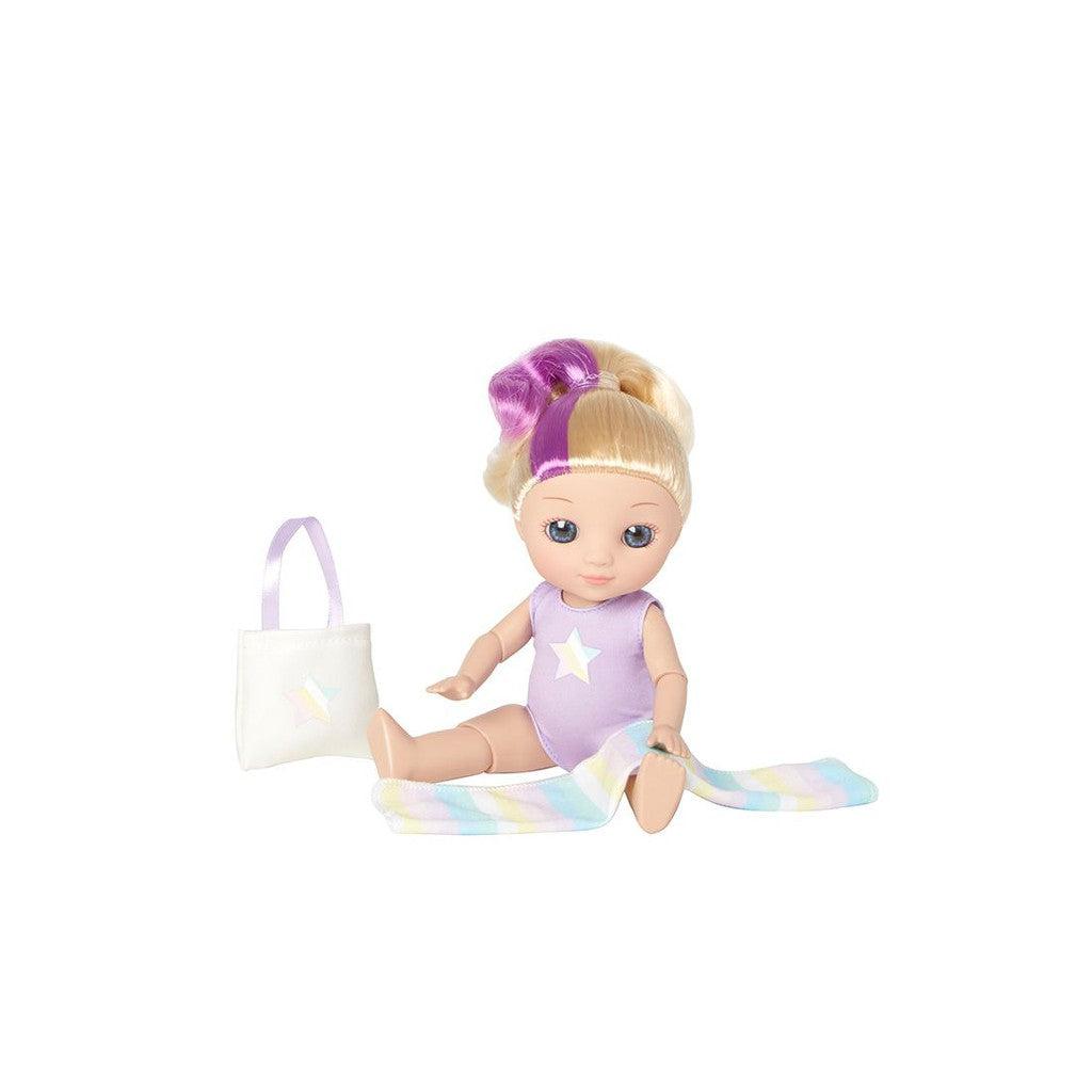 A doll with blonde hair, poised to dance in her vibrant purple outfit, sits beside a white bag and a rainbow-colored scarf.